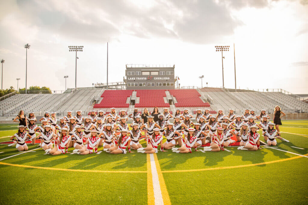 A group of cheerleaders smiles and poses together on a football field.