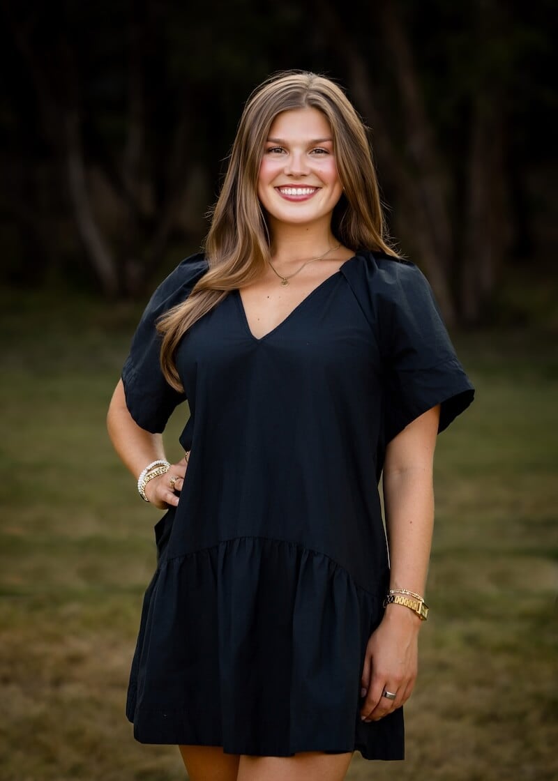 A woman with long brown hair poses in a black dress with one hand on her hip.