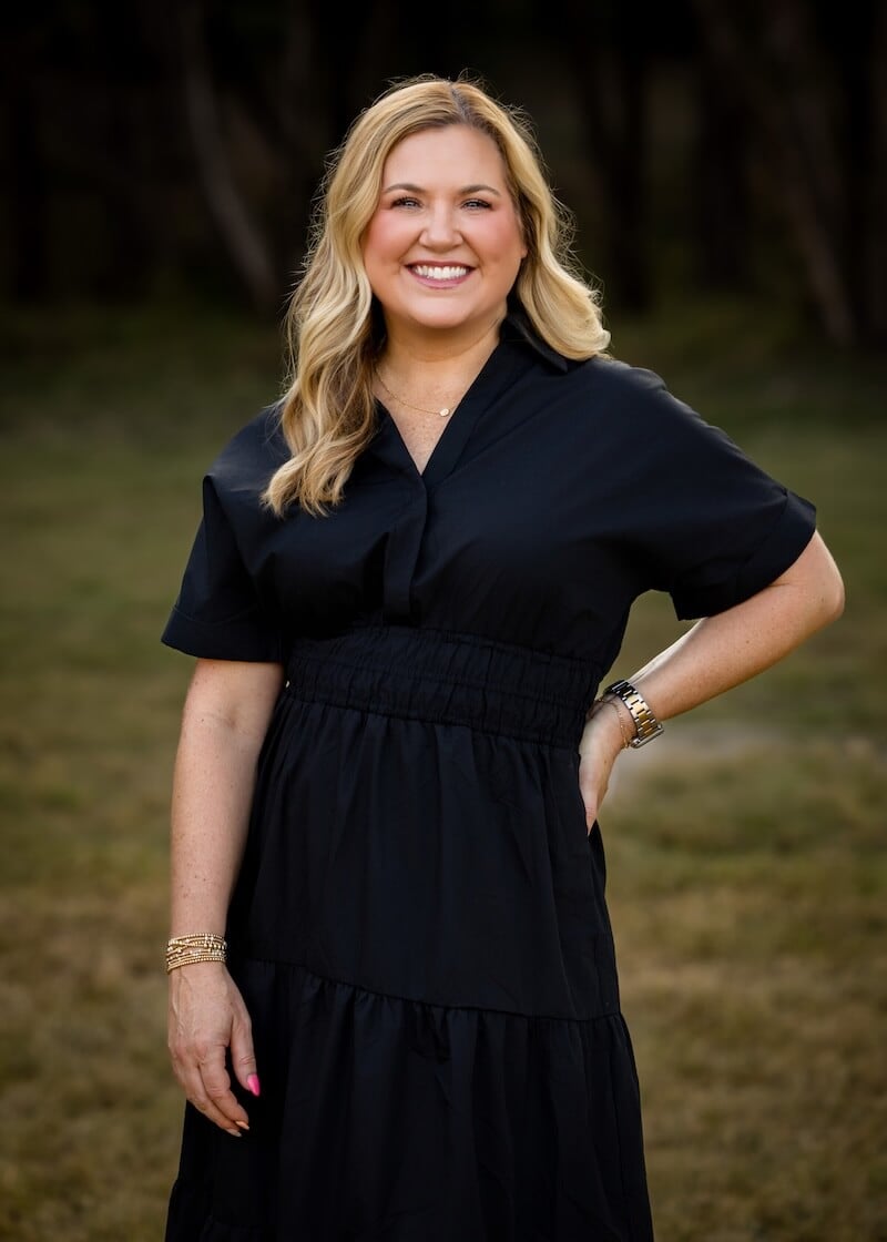 A blonde woman in a black dress poses with with one hand on her hip.