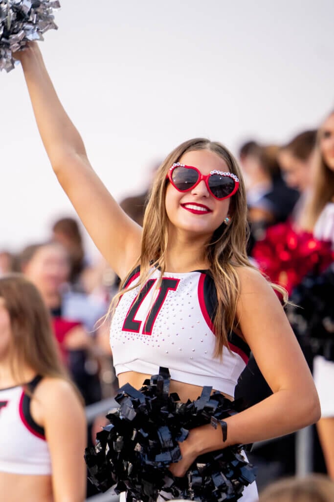 A smiling dance team member in red heart-shaped sunglasses and an “LT” uniform top raises a silver pom-pom overhead while cheering in the stands with teammates.