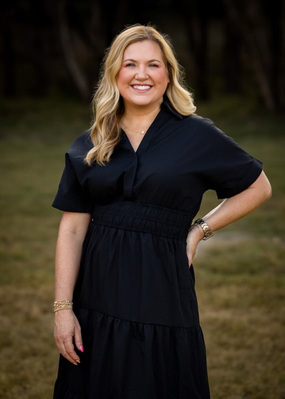 A blonde woman in a black dress poses with with one hand on her hip.
